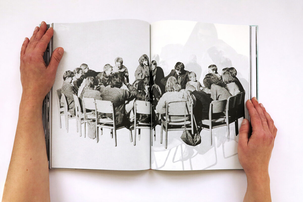 Aufgeschlagenes Fotobuch, das von zwei Händen aufgehalten wird. Im Buch zu sehen ist ein schwarzweiß-Foto einer großen Gruppe Frauen im Stuhlkreis, die sich angeregt unterhalten.