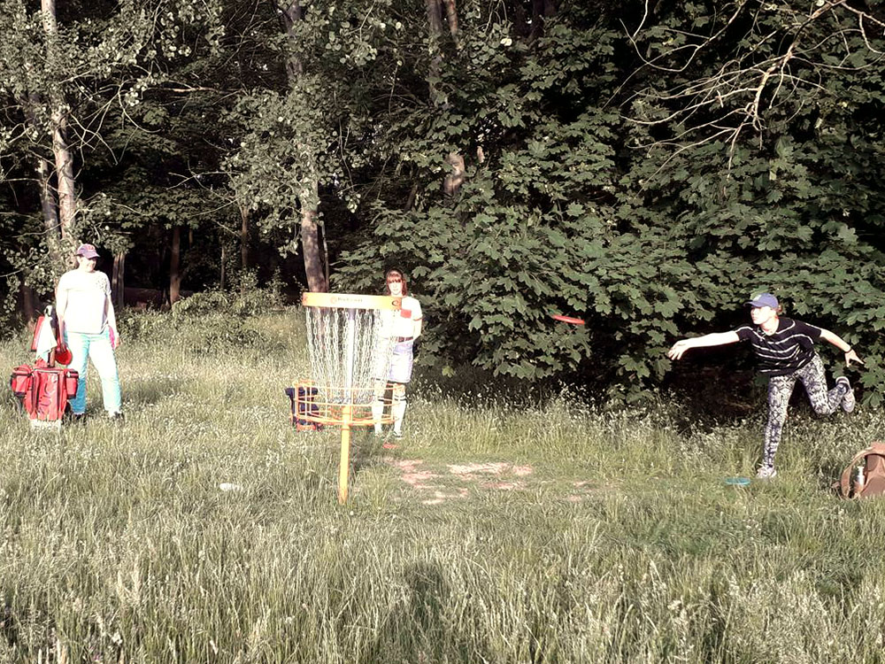 Eine Frau wirft eine Frisbee in einen Discgolf-Korb im Park, zwei weitere Frauen stehen daneben